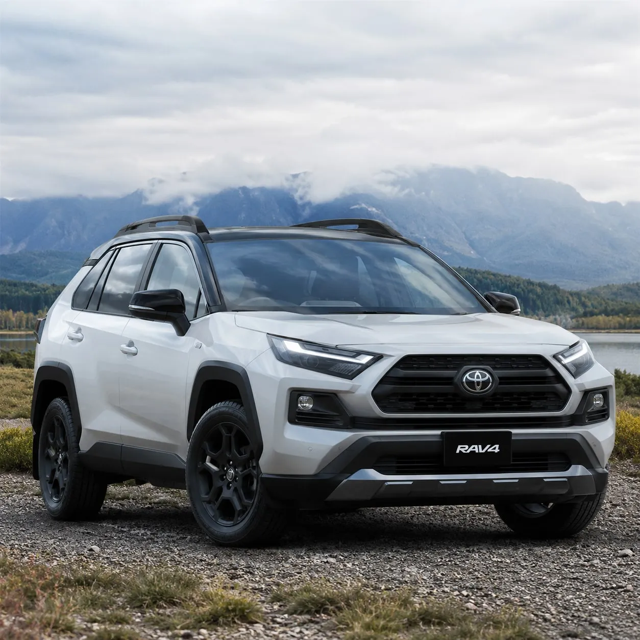 A silver SUV parked by a lake with mountains behind